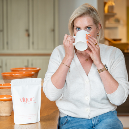 Woman drinking from a white mug in a kitchen setting with 'Vigor' products on the table.