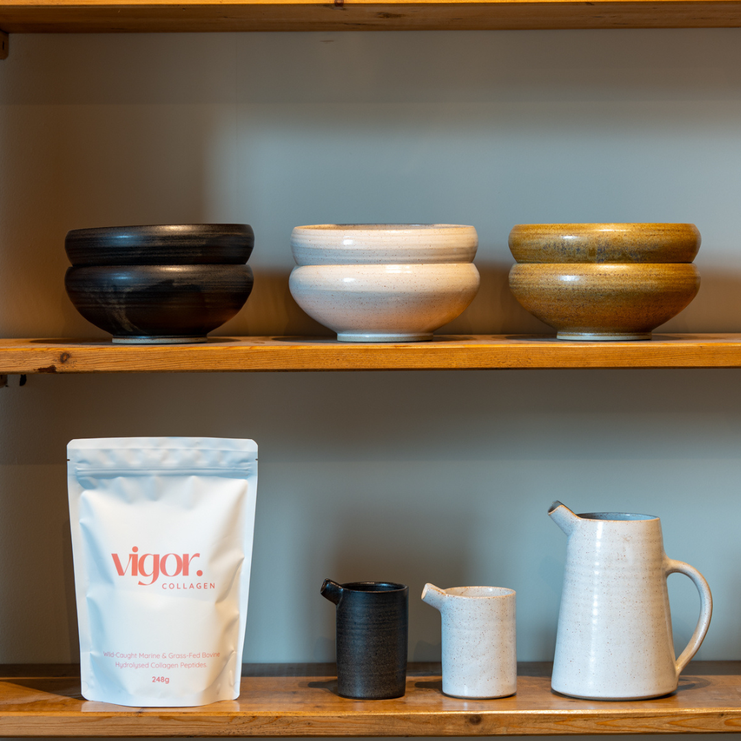 Vigor Collagen powder and set of ceramic bowls and mugs on a wooden shelf against a plain wall.