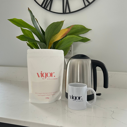 Vigor collagen powder bag and mug on a kitchen counter with a plant and kettle in the background.